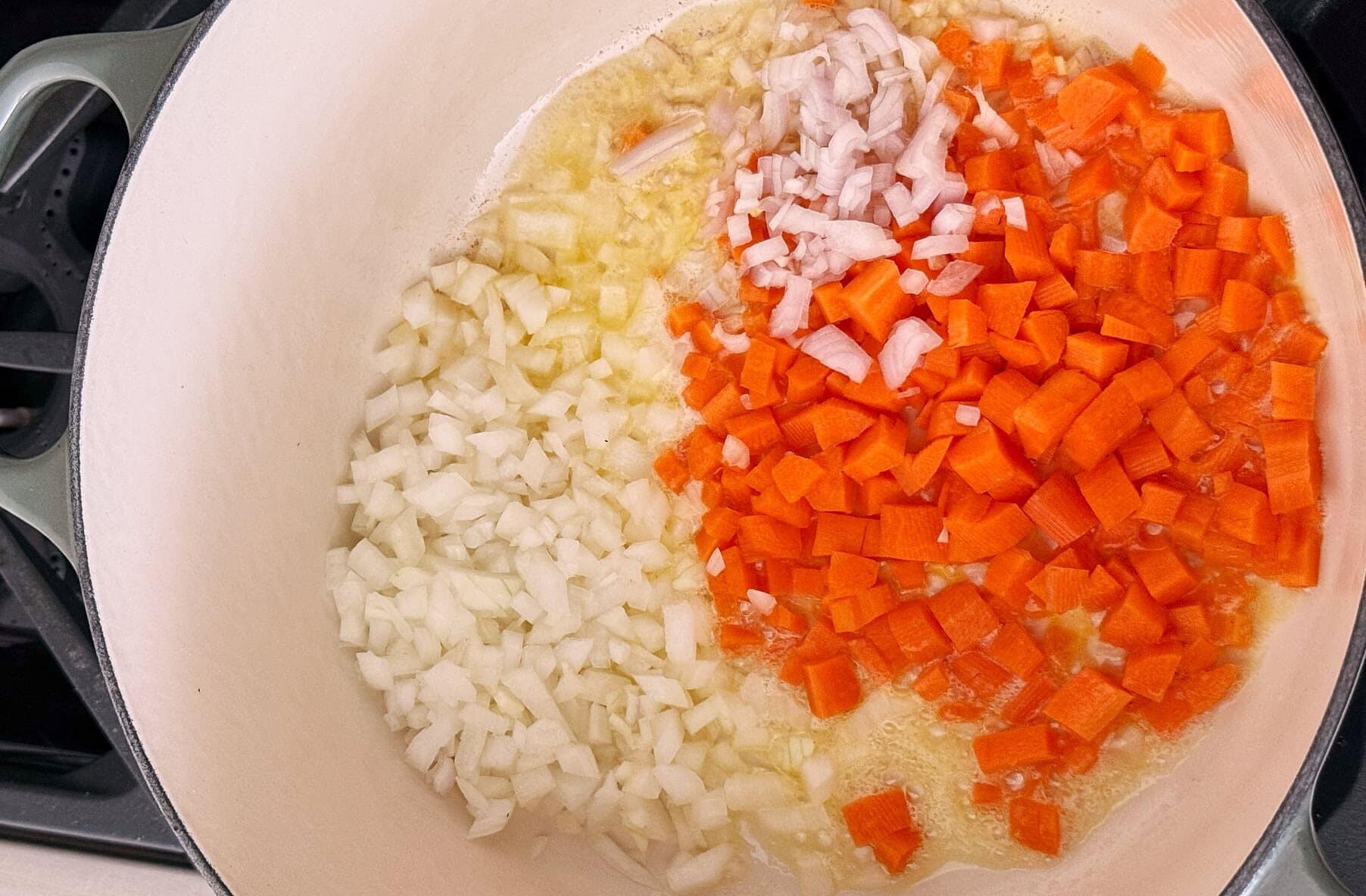 Diced onions, carrots, and shallots start sautéing in a large white pot on a stovetop.