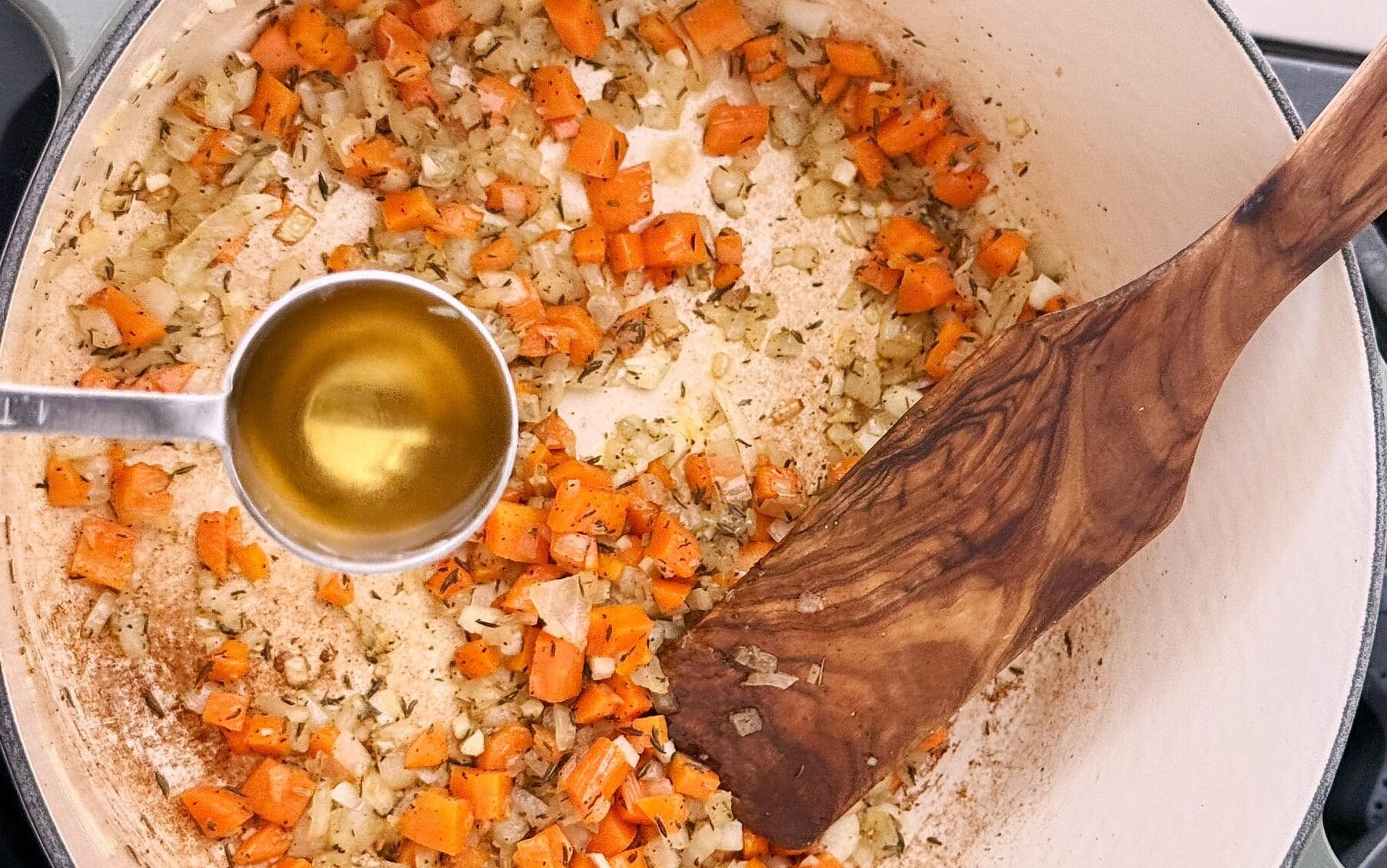 Diced carrots and onions are sautéed in a pot with a wooden spatula, while a metal measuring spoon holds liquid above the mixture.