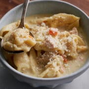 A bowl of creamy chicken and tortellini soup topped with grated cheese and black pepper, with a spoon lifting a portion.