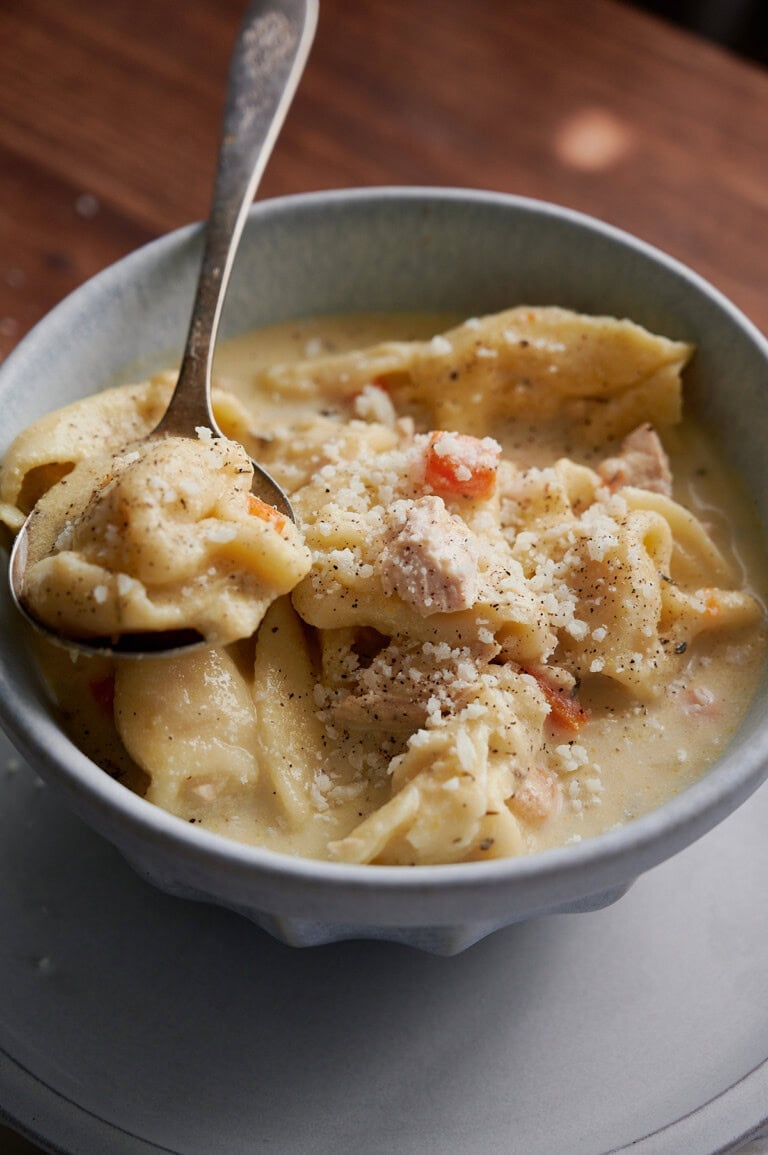 A bowl of creamy chicken and tortellini soup topped with grated cheese and black pepper, with a spoon lifting a portion.