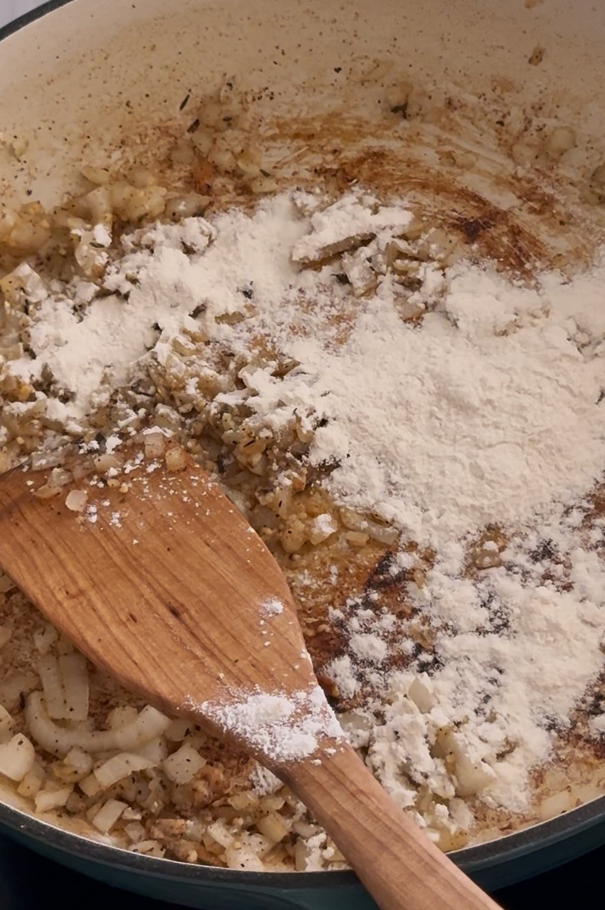 Chopped onions sautéing in a pan with flour sprinkled on top and a wooden spatula stirring the mixture.