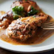 A plate with two pieces of cooked chicken in a brown sauce, garnished with herbs, and a fork resting on the side.