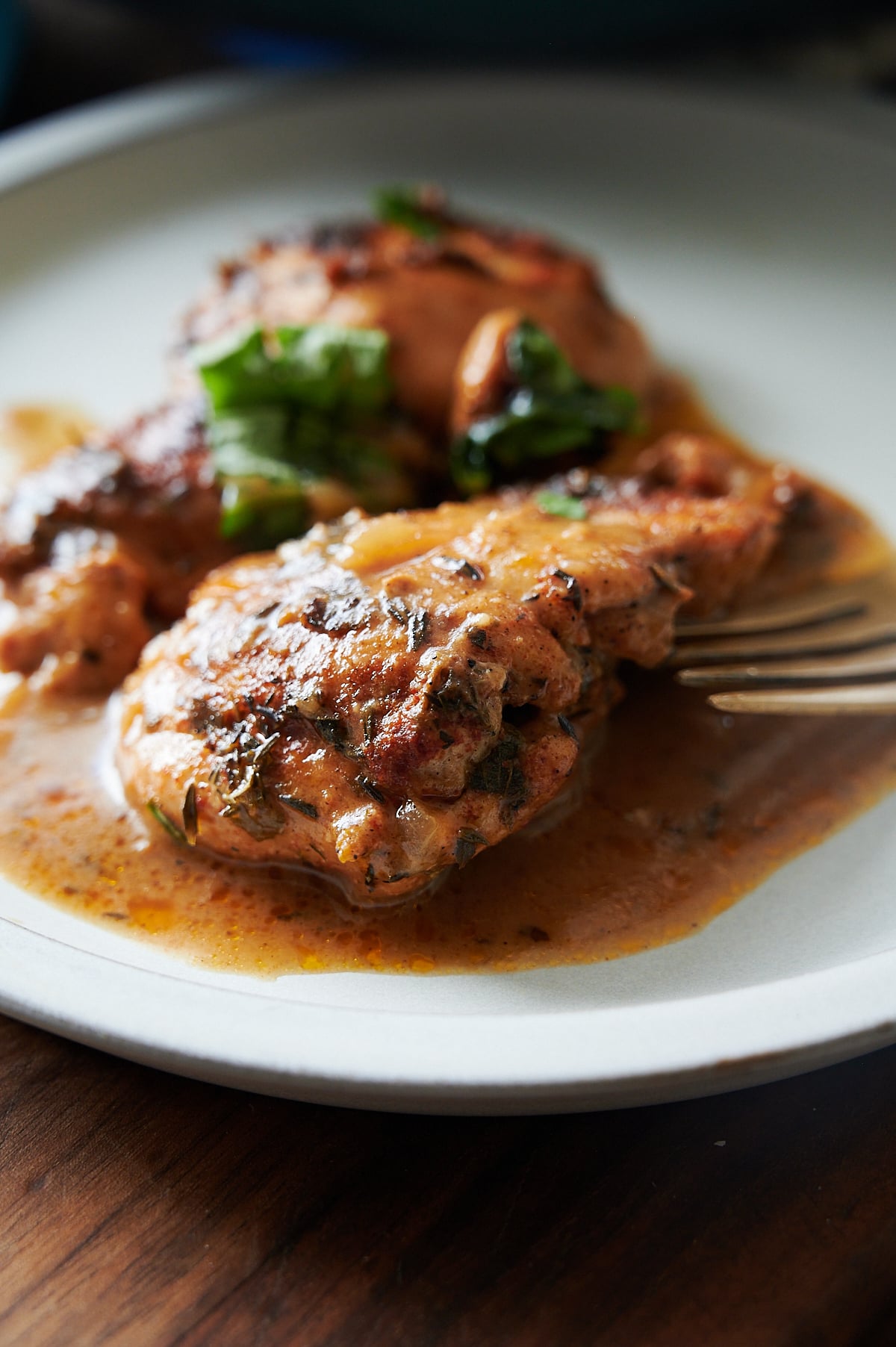 A plate with two pieces of cooked chicken in a brown sauce, garnished with herbs, and a fork resting on the side.