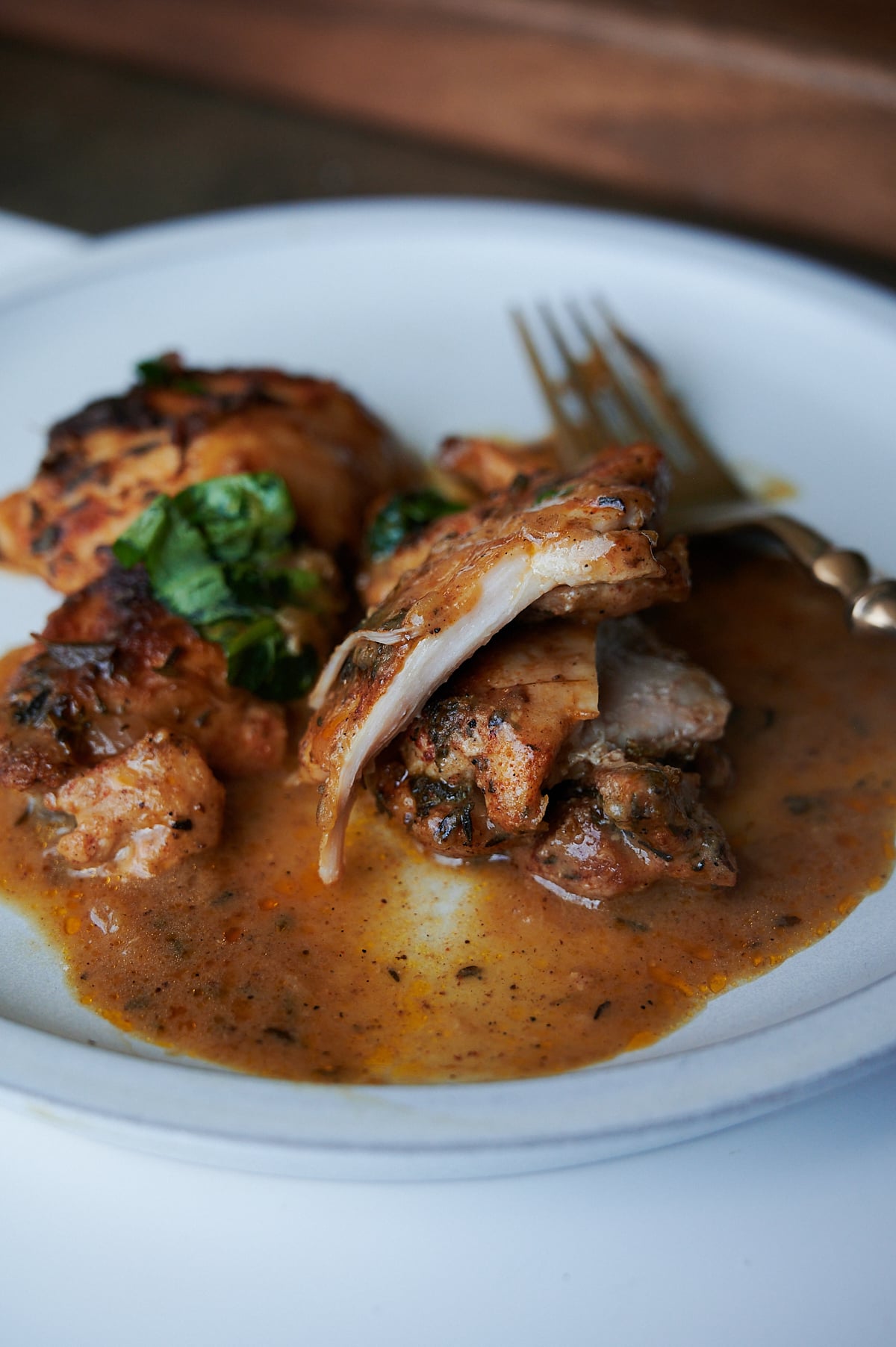 A plate of cooked chicken pieces with brown sauce, garnished with herbs, next to a fork on a white plate.