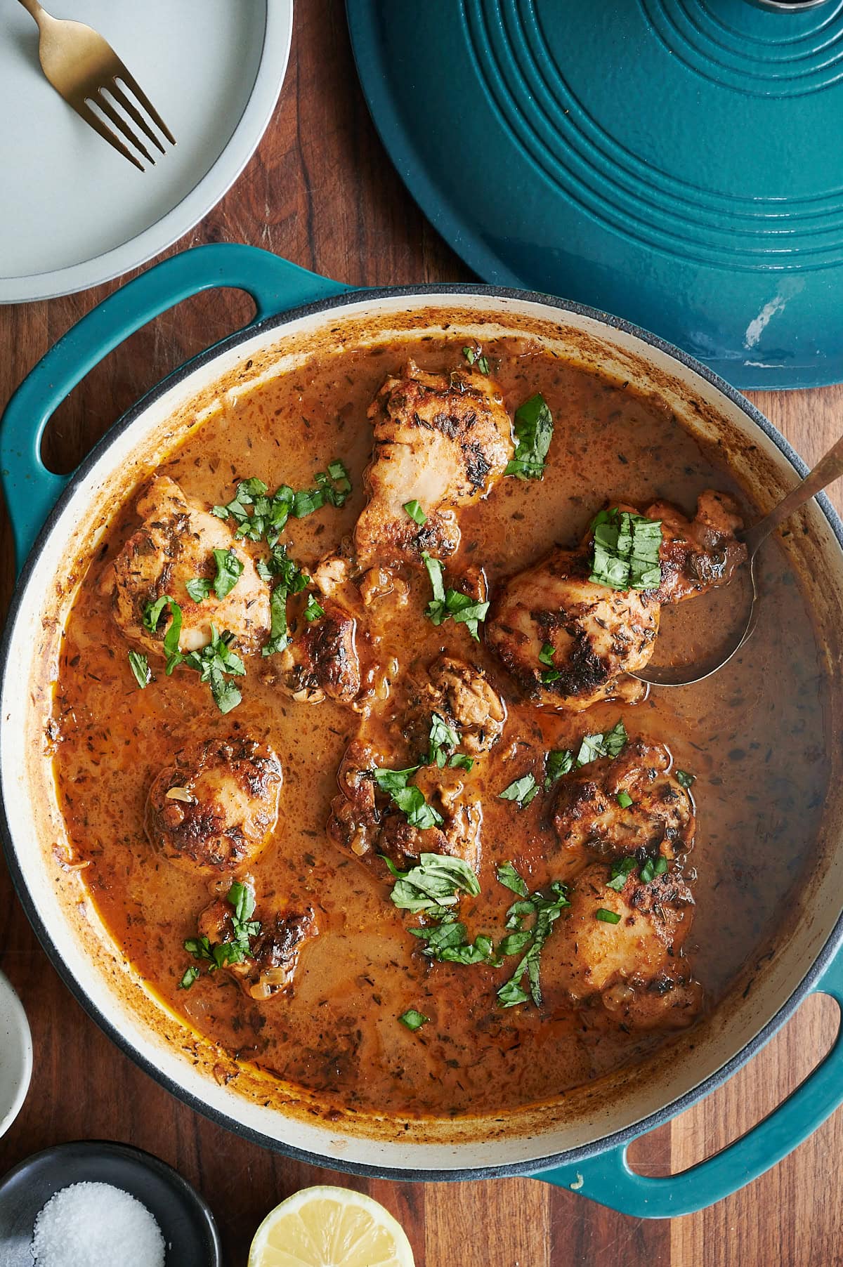 A round teal Dutch oven filled with cooked, saucy chicken pieces garnished with chopped herbs, sits on a wooden table with a serving spoon, lemon, and salt nearby.