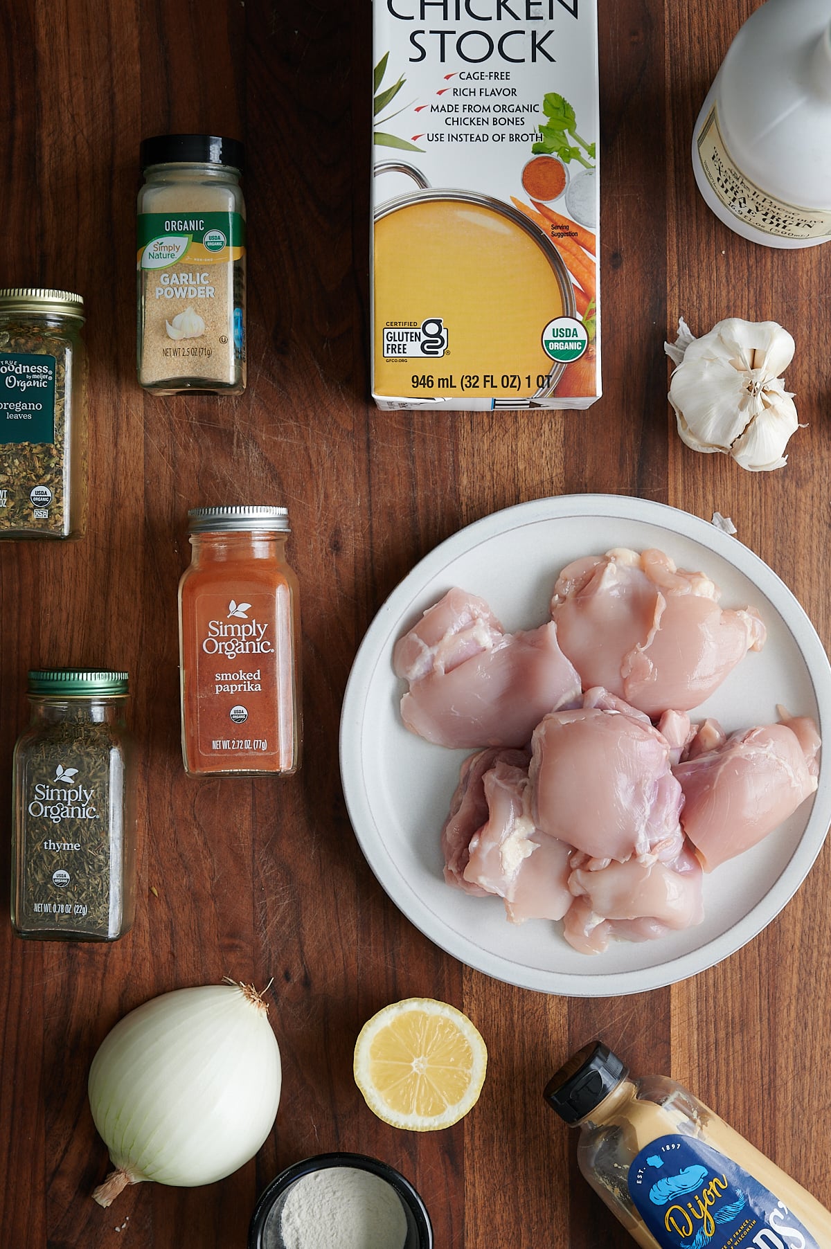 Assorted ingredients on a wooden surface, including raw chicken thighs on a plate, chicken stock, spices, garlic, onion, lemon, Dijon mustard, and a bottle of olive oil.