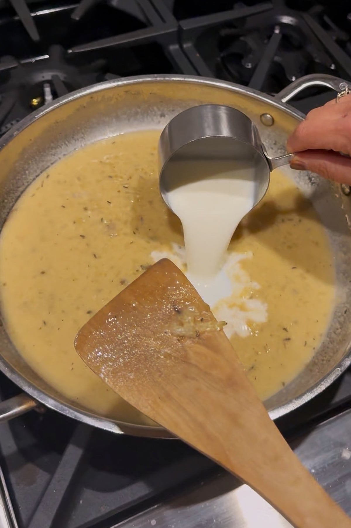 A hand pours milk from a metal measuring cup into a skillet with a creamy sauce on a stove, next to a wooden spatula.