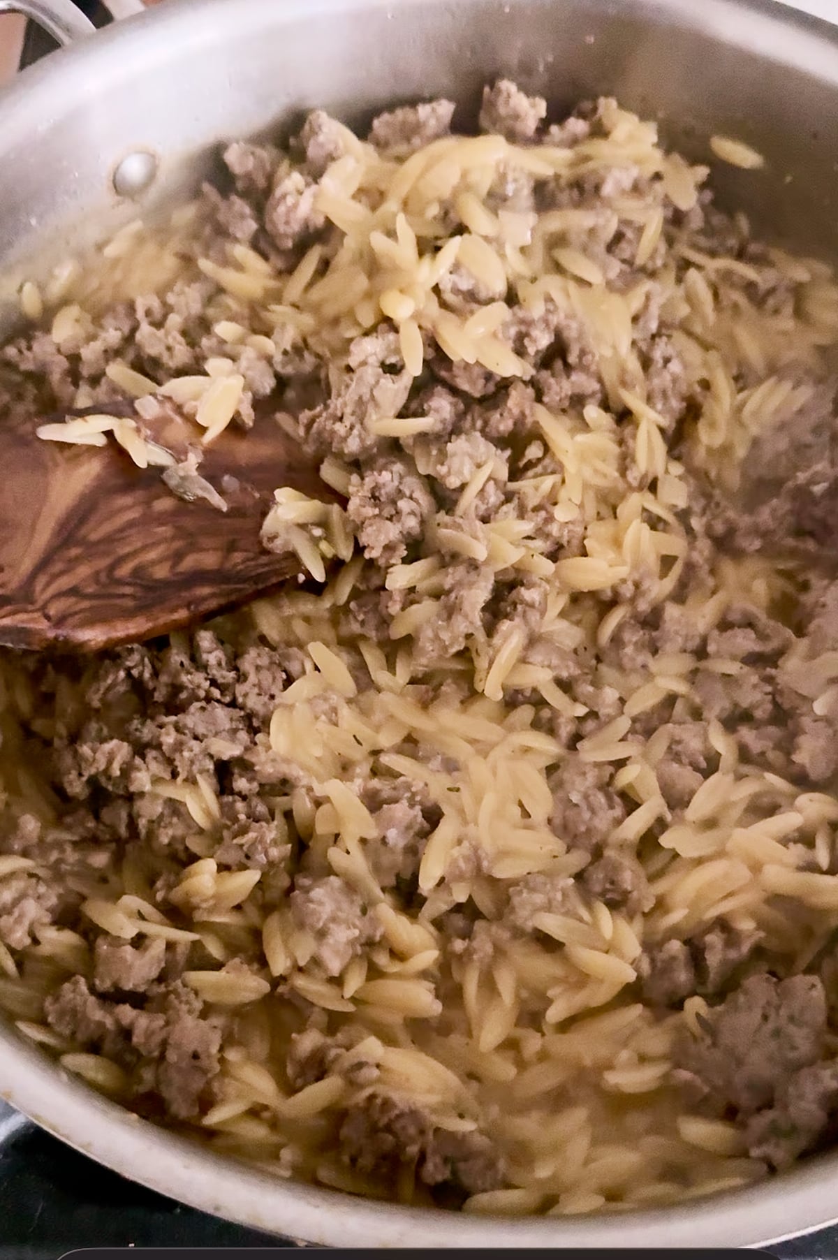 A close-up of a pot containing cooked orzo pasta and Italian sausage being stirred with a wooden spoon.