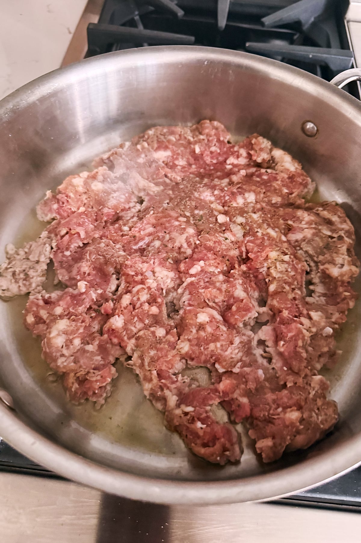 Ground Italian sausage cooking in a stainless steel skillet on a stovetop, starting to brown around the edges.