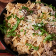 A red skillet filled with orzo pasta, ground sausage, grated cheese, and chopped fresh herbs, garnished with lemon zest.