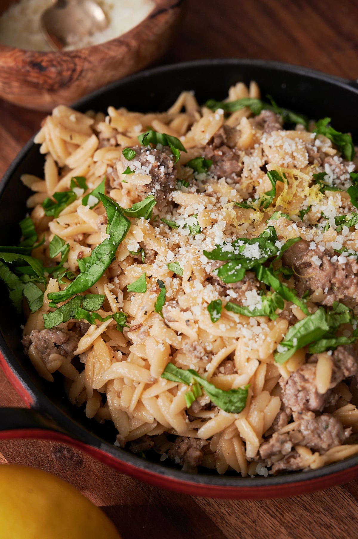 A red skillet filled with orzo pasta, ground sausage, grated cheese, and chopped fresh herbs, garnished with lemon zest.