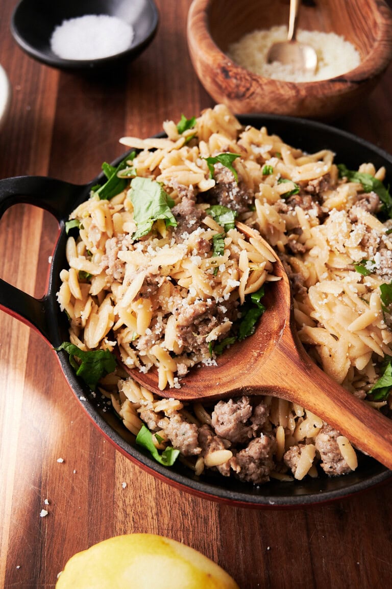 A skillet of orzo pasta with ground sausage, spinach, and grated cheese, served with a wooden spoon on a wooden table.