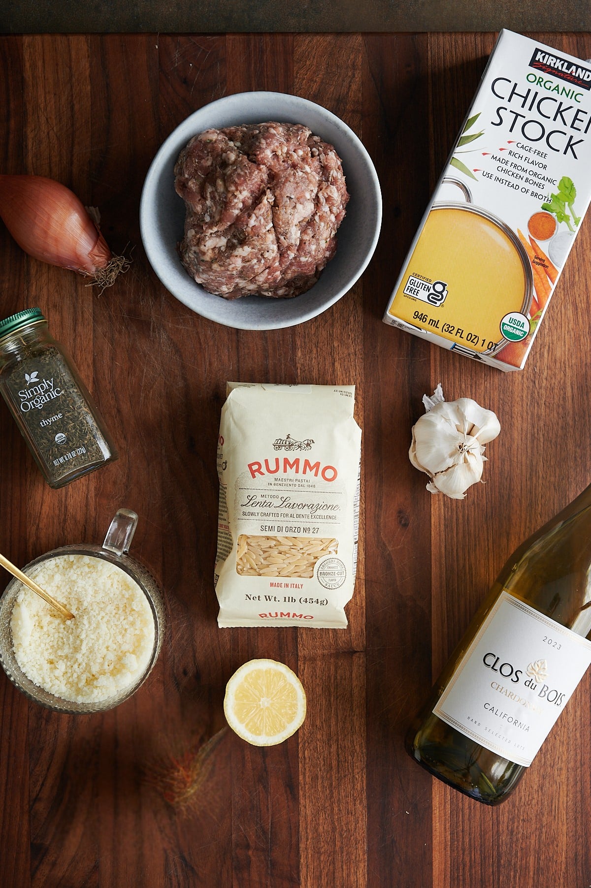 Raw ground sausage, chicken stock, orzo, garlic, shallot, onion, dried thyme, grated cheese, a lemon half, and a bottle of white wine on a wooden board.