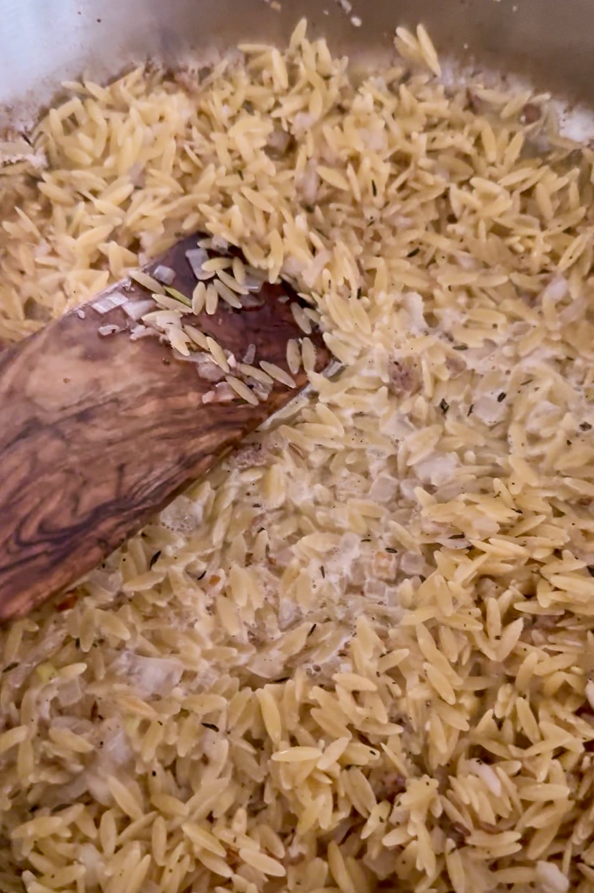 A wooden spoon stirring orzo pasta in a pan with visible liquid and seasonings.