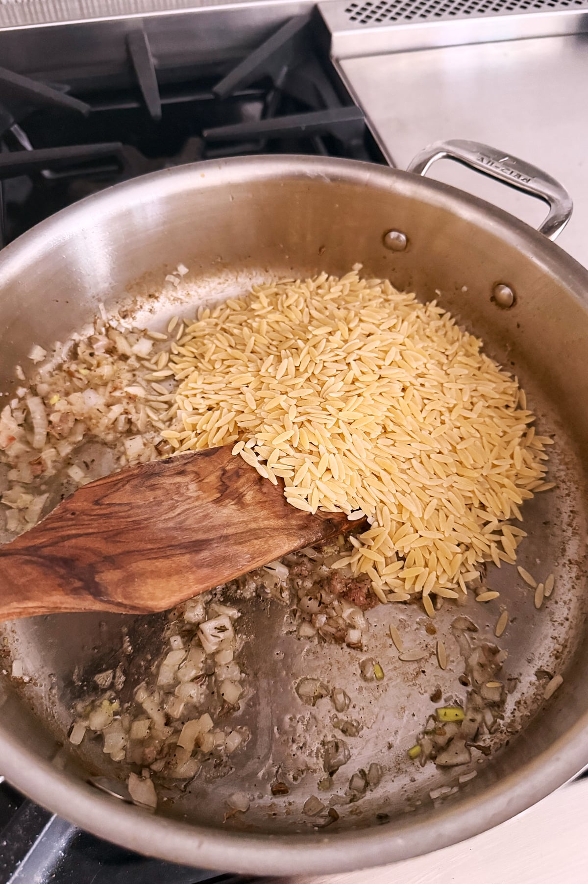 Orzo pasta is added to sautéed shallots in a stainless steel pan on a stovetop, with a wooden spatula stirring the mixture.
