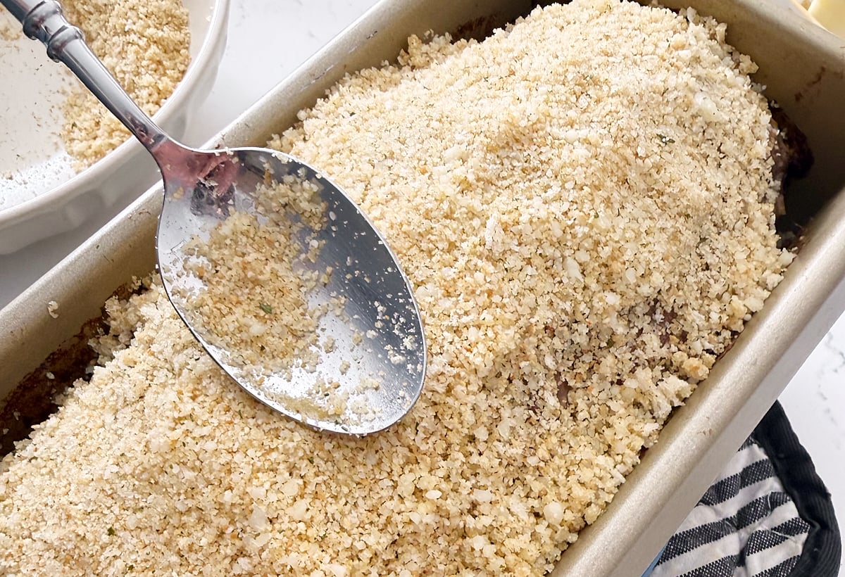 A metal spoon spreads a layer of seasoned breadcrumbs over a meatloaf in a baking pan, preparing it for baking.