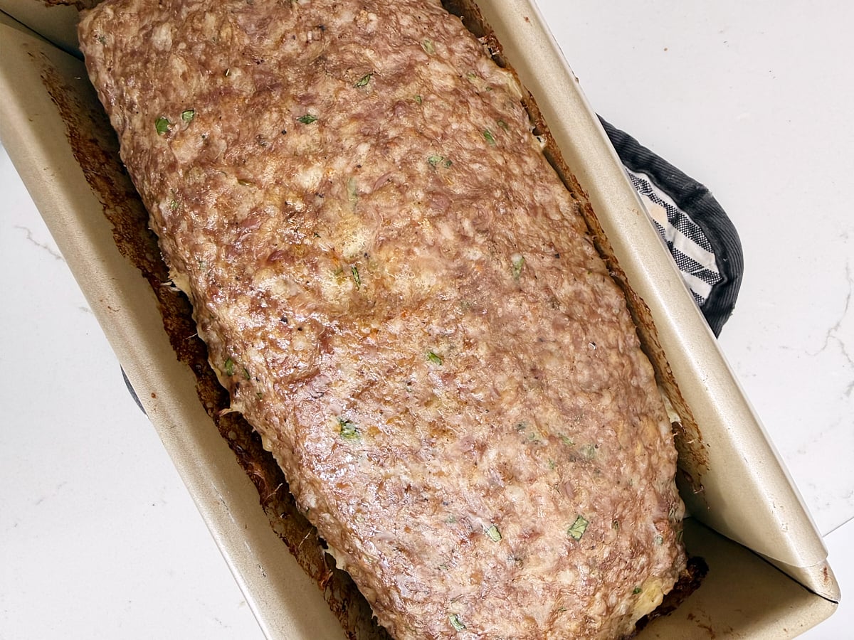 A cooked meatloaf rests in a rectangular baking pan on a white surface, with a black and white oven mitt partially visible.