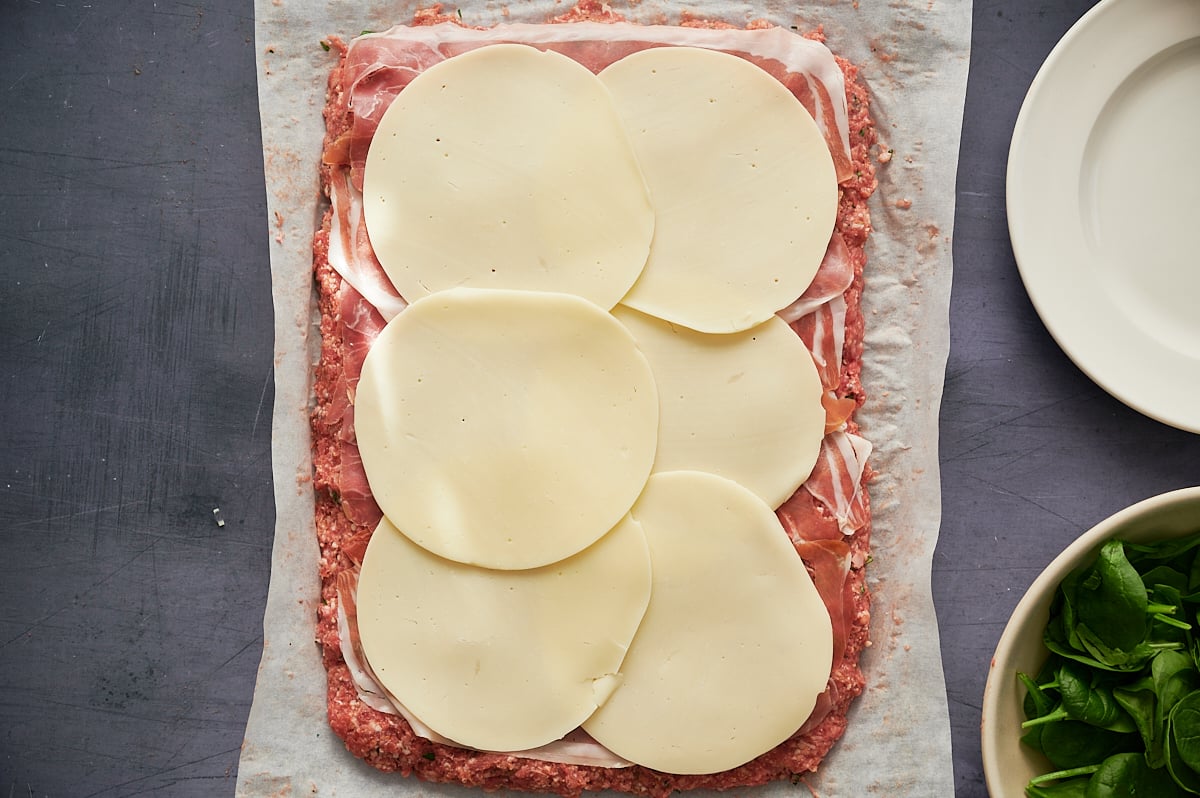 Rectangular layer of meatloaf mix topped with sliced ham and six overlapping slices of provolone cheese on parchment paper, next to empty and spinach-filled bowls.