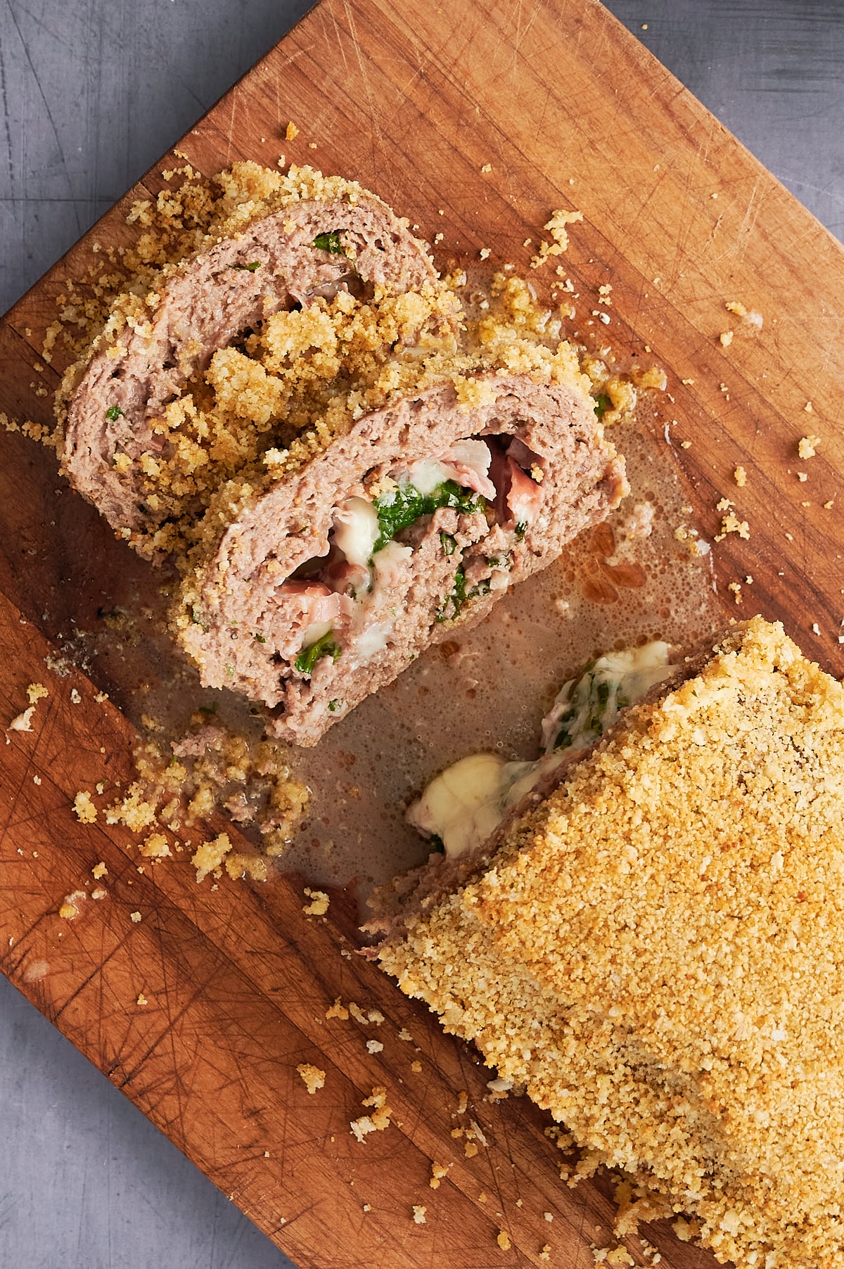 A sliced meatloaf stuffed with cheese, spinach, and ham sits on a wooden cutting board, topped with a layer of breadcrumbs.