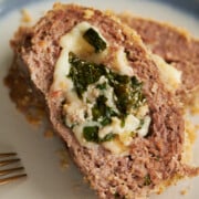 A close-up of a sliced meatloaf stuffed with melted cheese and spinach on a light-colored blue-rimmed plate.