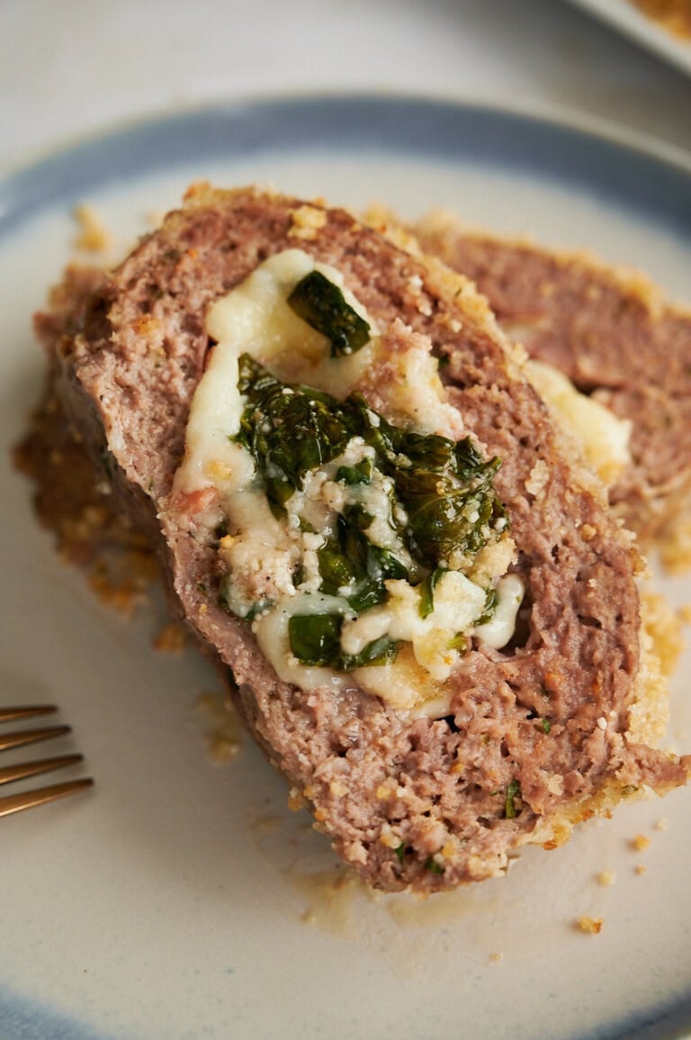A close-up of a sliced meatloaf stuffed with melted cheese and spinach on a light-colored blue-rimmed plate.
