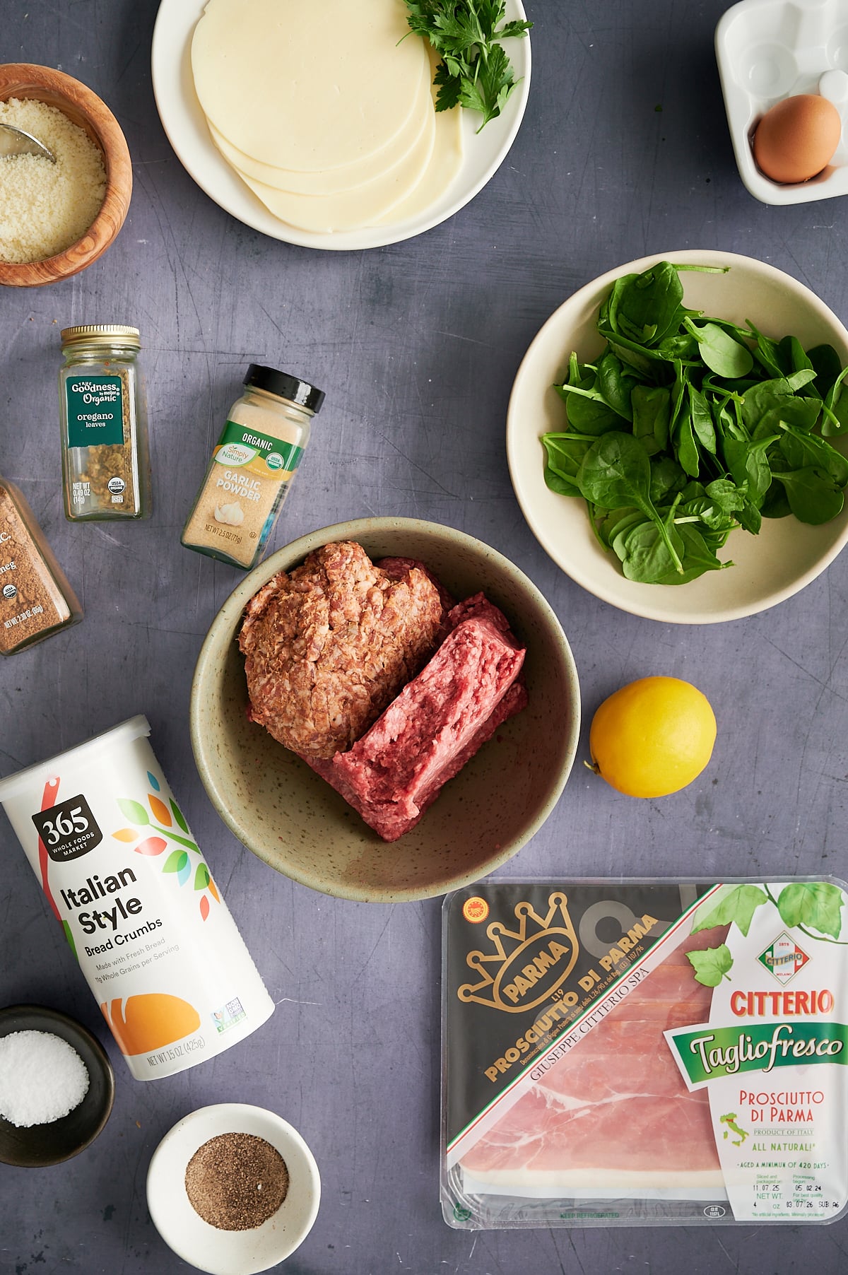 Meatloaf ingredients arranged on a gray surface, including ground meat, sliced cheese, spinach, lemon, prosciutto, bread crumbs, seasonings, salt, pepper, and parsley.