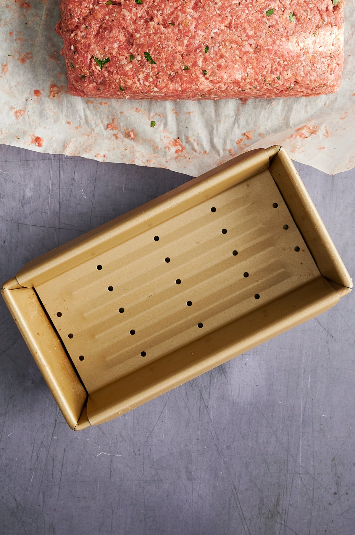An empty gold loaf pan with holes sits on a gray surface next to a sheet of parchment paper holding a portion of meatloaf mixture.