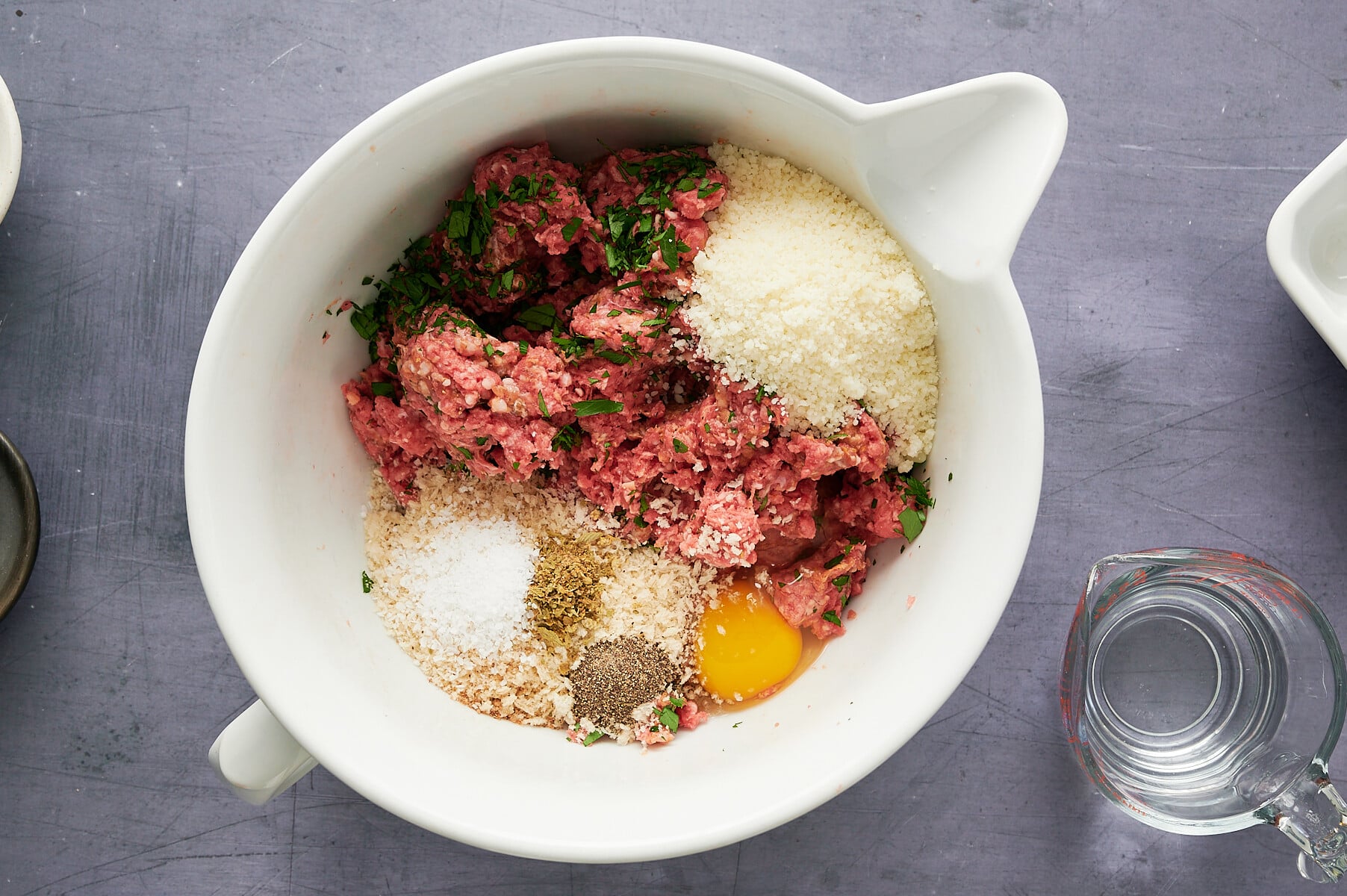 A white mixing bowl with ground meat, an egg, breadcrumbs, grated cheese, salt, pepper, and fresh herbs, ready to be mixed.