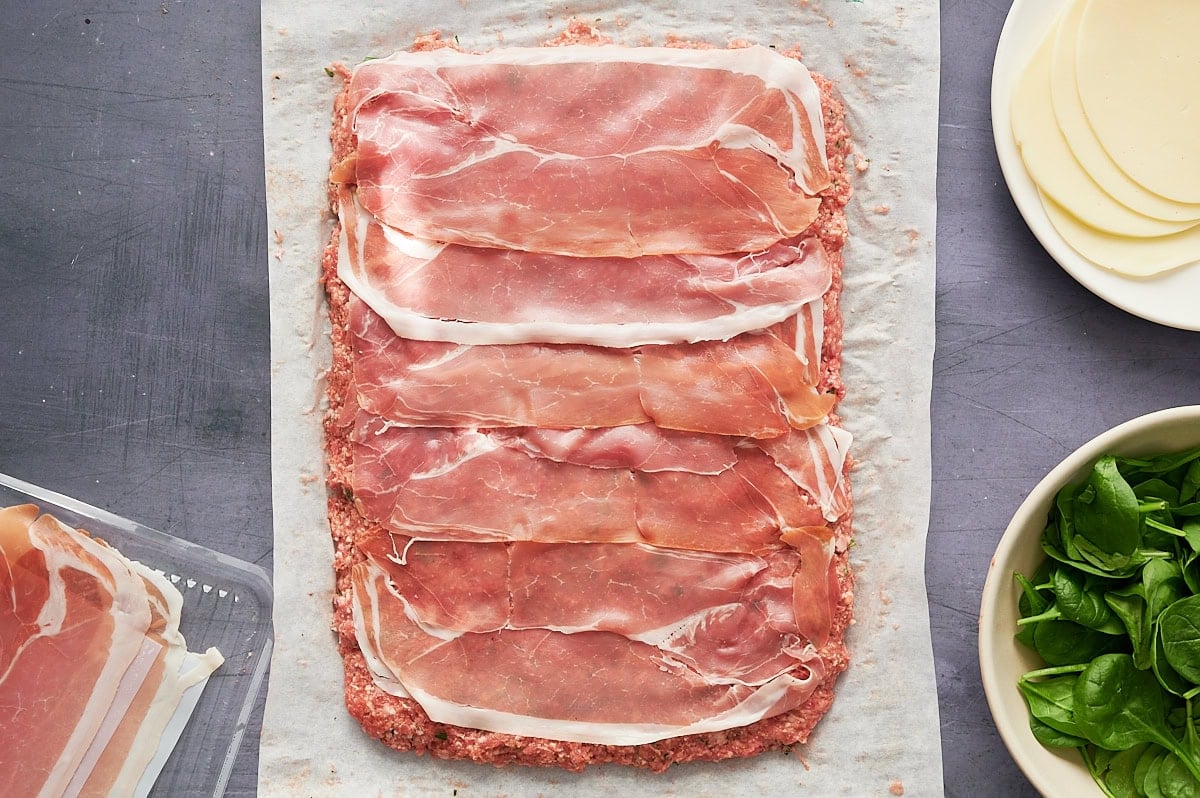 Layered slices of prosciutto on top of meatloaf mix spread on parchment paper, with a bowl of spinach and slices of cheese nearby.