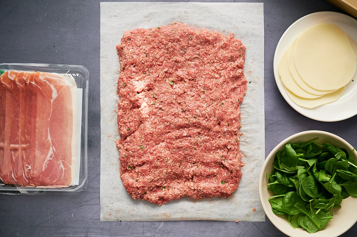 Raw meatloaf mix spread on parchment paper, with a bowl of spinach, a plate of cheese slices, and a package of sliced prosciutto arranged nearby on a gray surface.