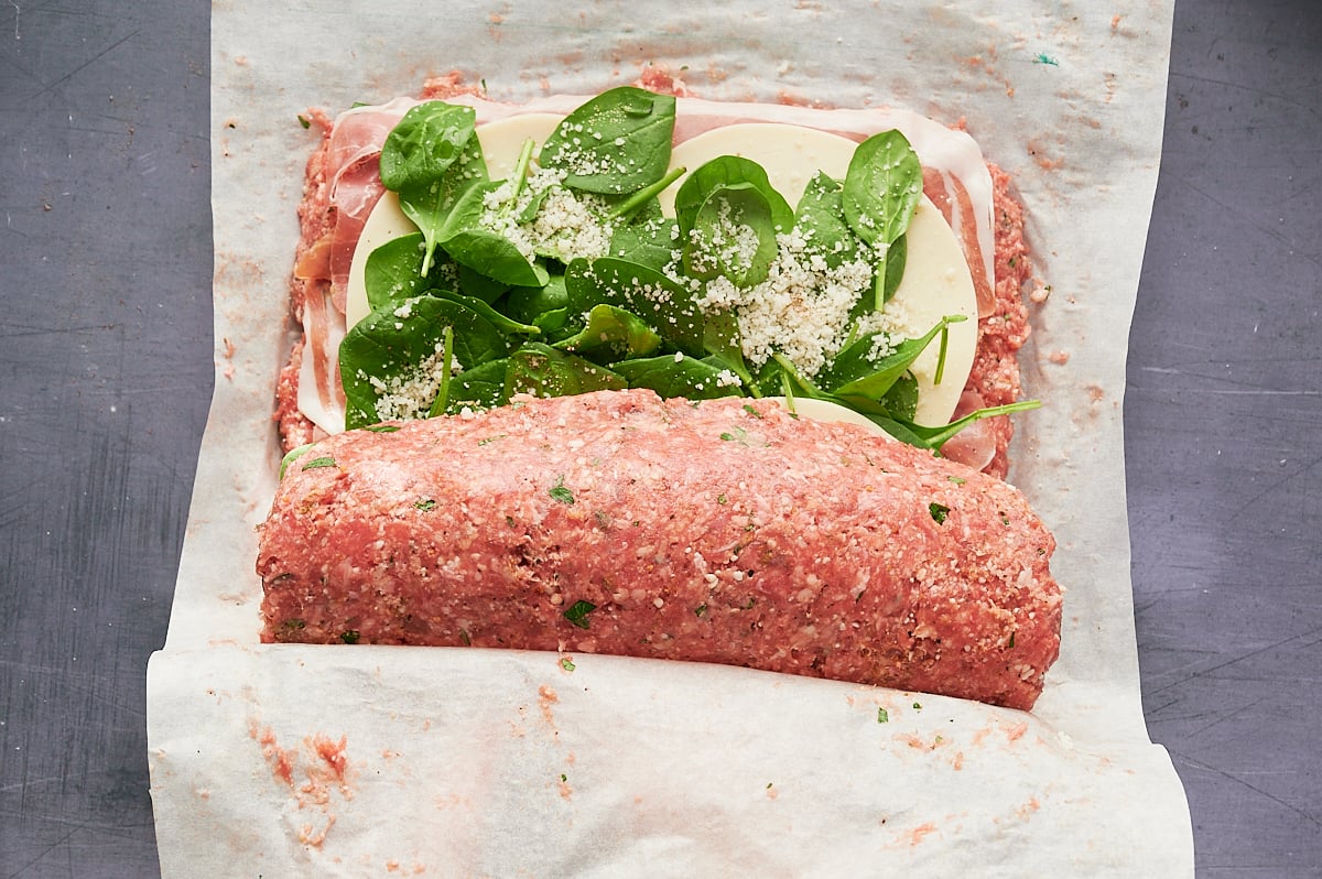 Raw meatloaf mixture being rolled around layers of sliced cheese, prosciutto, fresh spinach, and grated cheese on parchment paper.