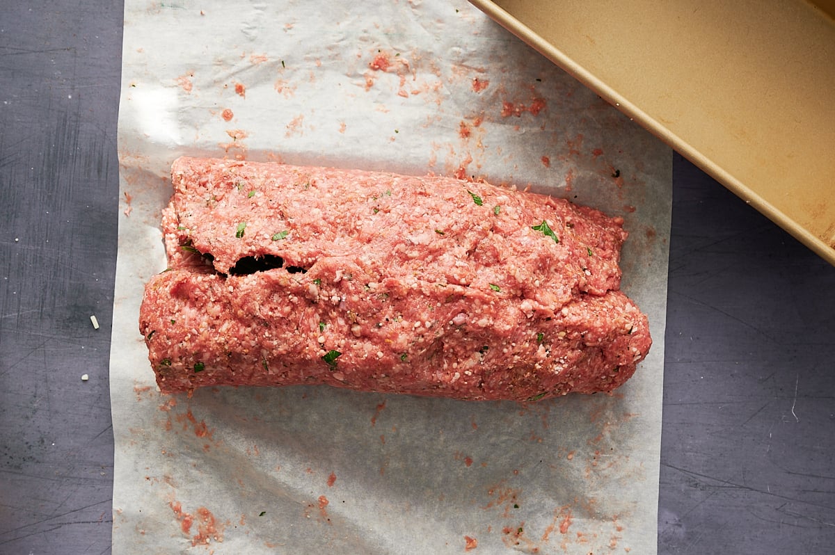 A loaf-shaped portion of raw ground meat stuffed with meat, cheese and herbs and seasoning sits on a sheet of parchment paper next to a baking dish.