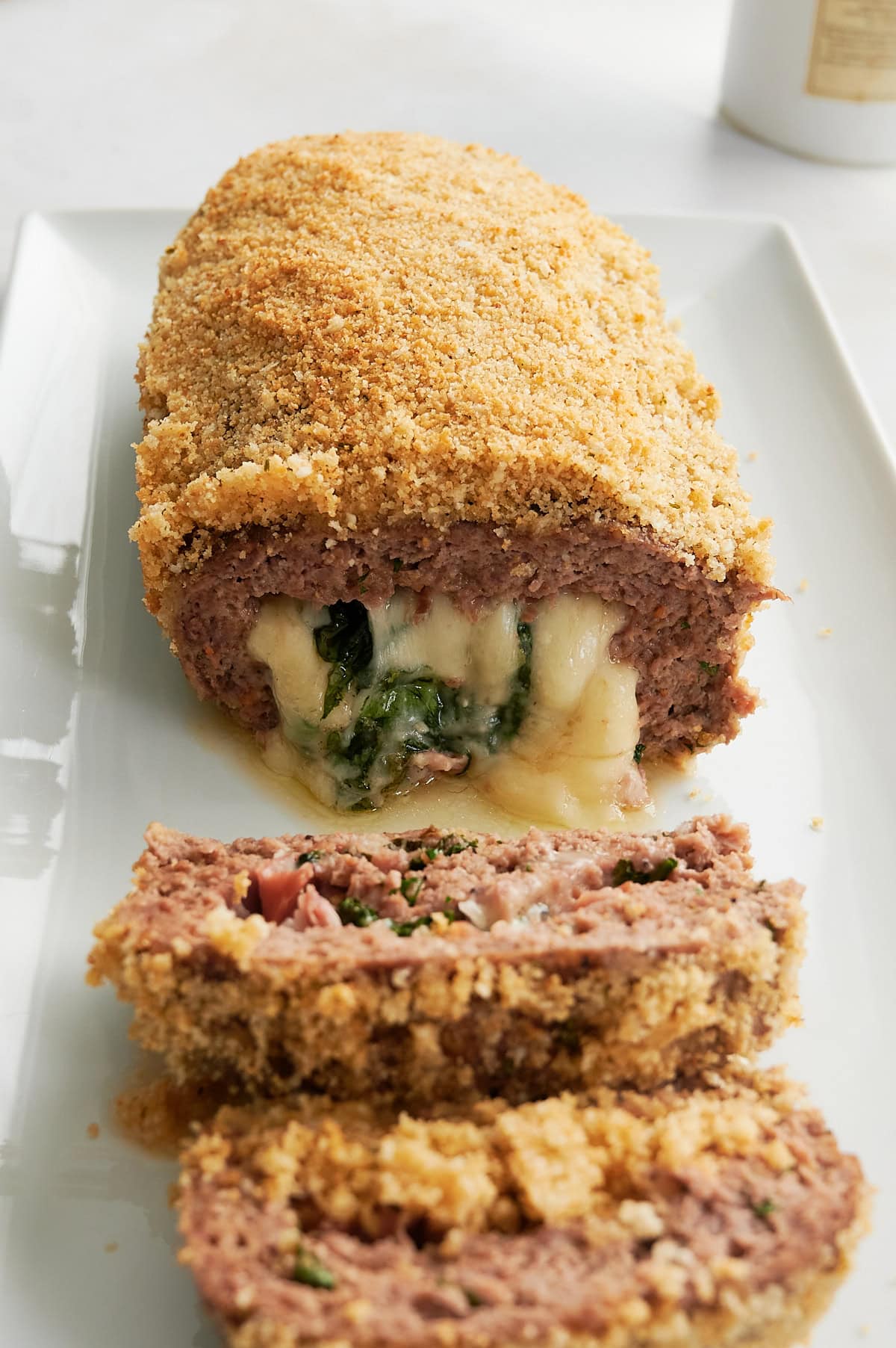 A breadcrumb topped meatloaf stuffed with melted cheese, prosciutto and spinach sliced on a white rectangular platter.