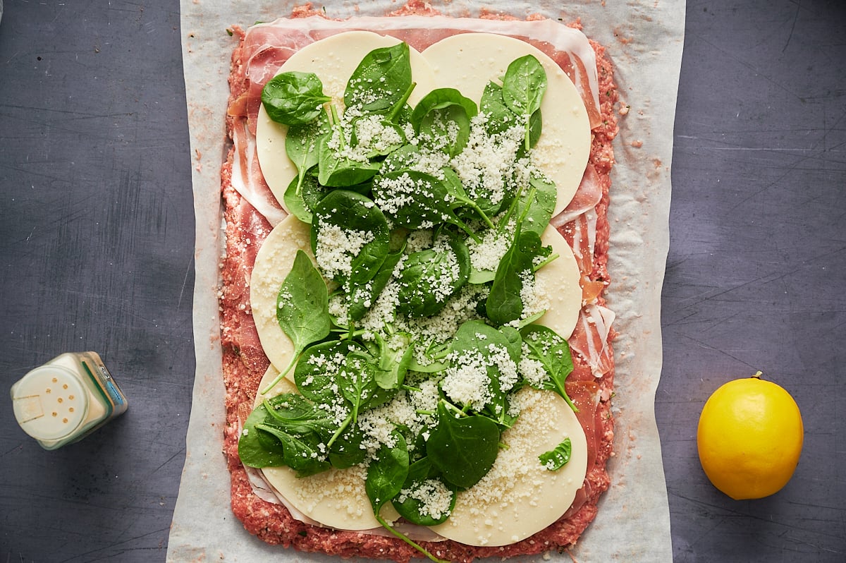 Rectangular layer of ground meat topped with sliced cheese, spinach leaves, grated cheese, and slices of deli meat on parchment paper, with a lemon and seasoning shaker beside it.