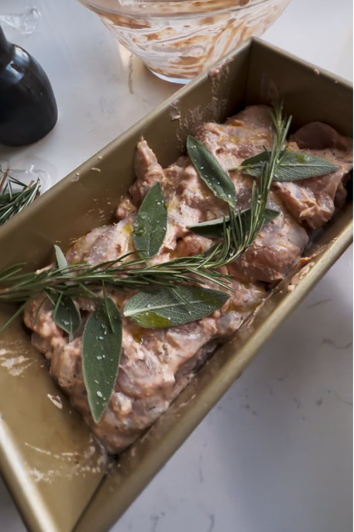 Raw marinated chicken thighs in a loaf pan topped with fresh rosemary and sage leaves, ready for cooking. A mixing bowl and pepper grinder are nearby on the countertop.