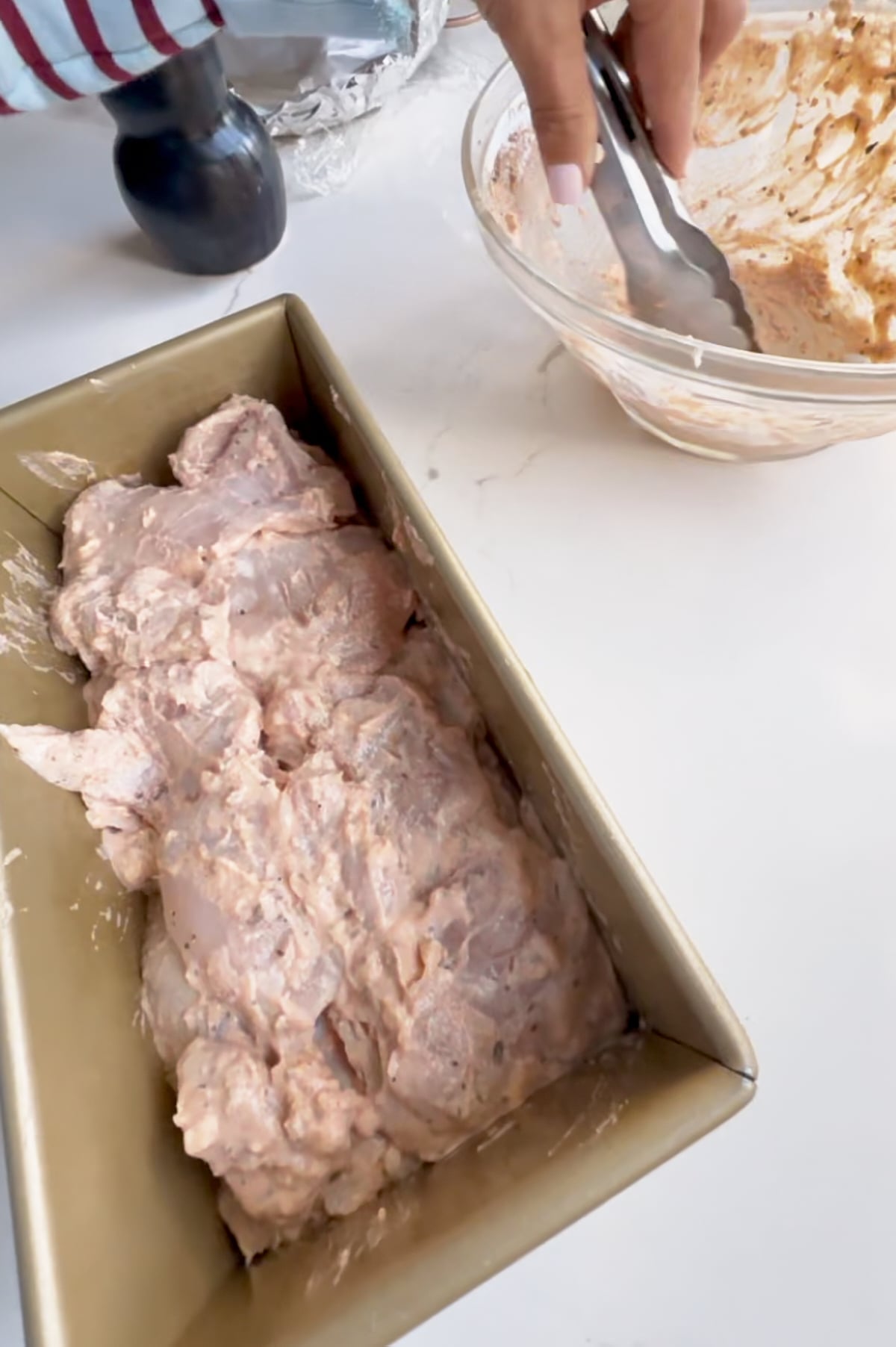 Raw, marinated Italian loaf pan chicken is arranged in a pan, as a person uses tongs beside a glass bowl of marinade on a white countertop.