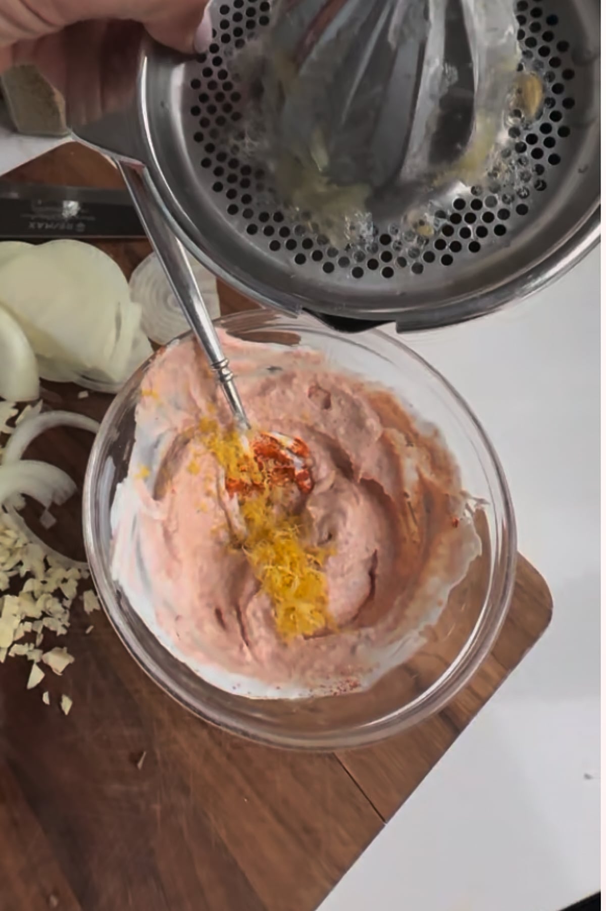 A hand squeezes a citrus juicer over a bowl of creamy mixture with orange zest on top, next to chopped onions and garlic on a wooden cutting board.