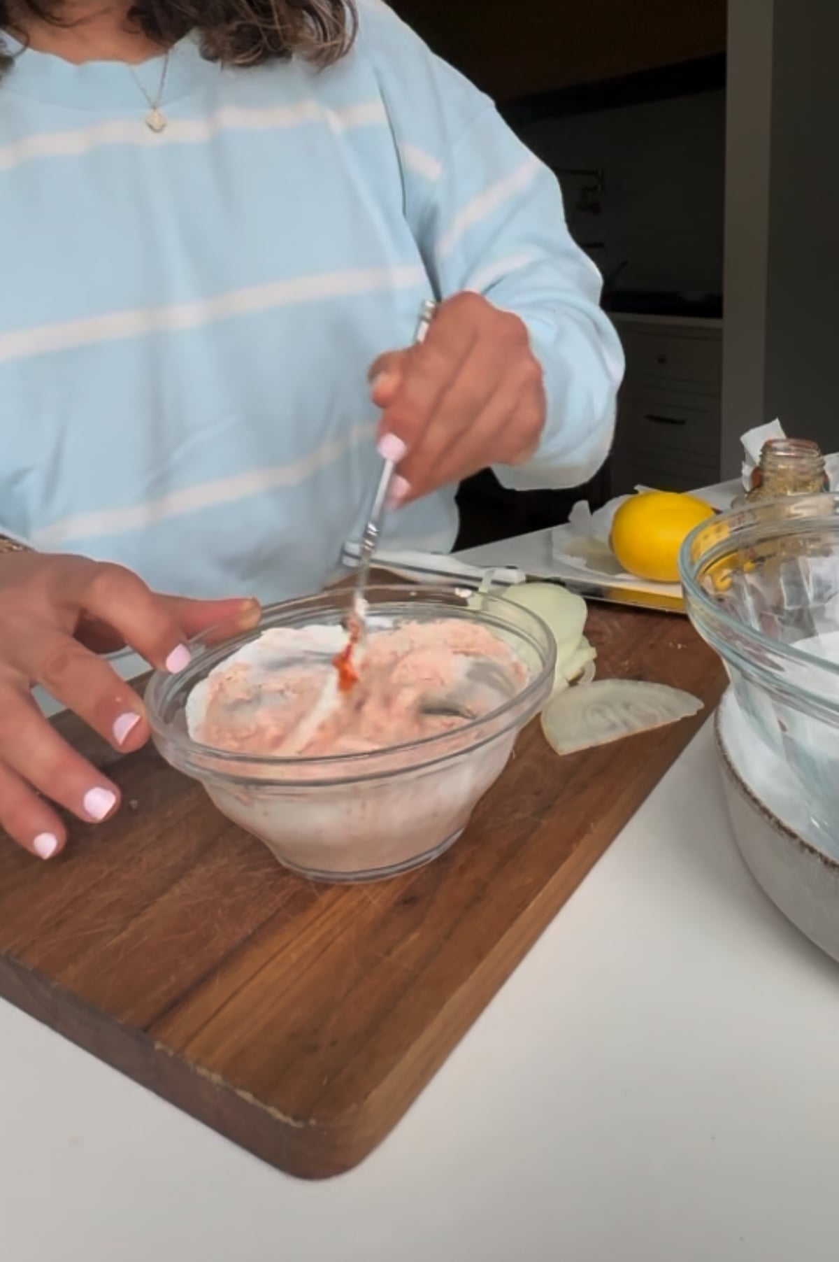 A person stirring a pink mixture in a glass bowl on a wooden cutting board with sliced onions and a lemon nearby.