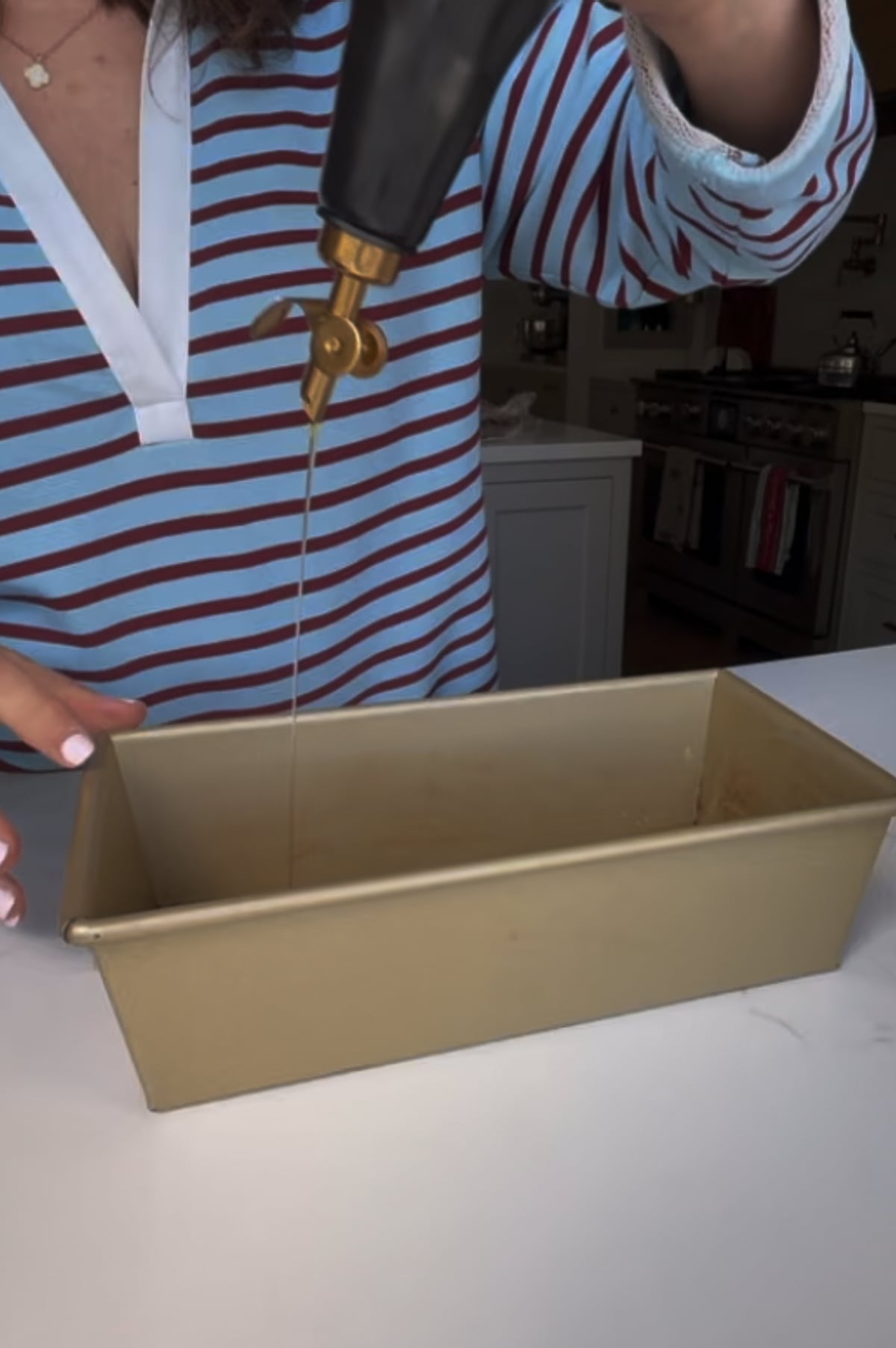 A person in a striped shirt pours oil from a bottle into a rectangular baking pan on a kitchen counter.