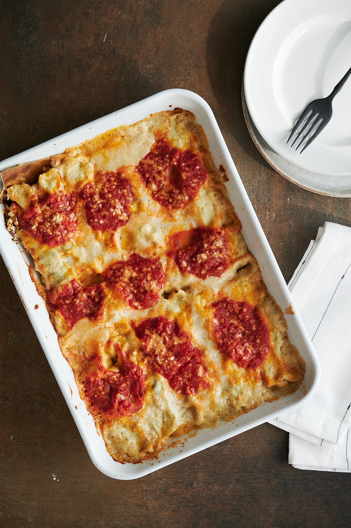 Baked chicken cannelloni in a white rectangular dish, topped with tomato sauce and cheese, next to stacked white plates, a fork, and a folded napkin.