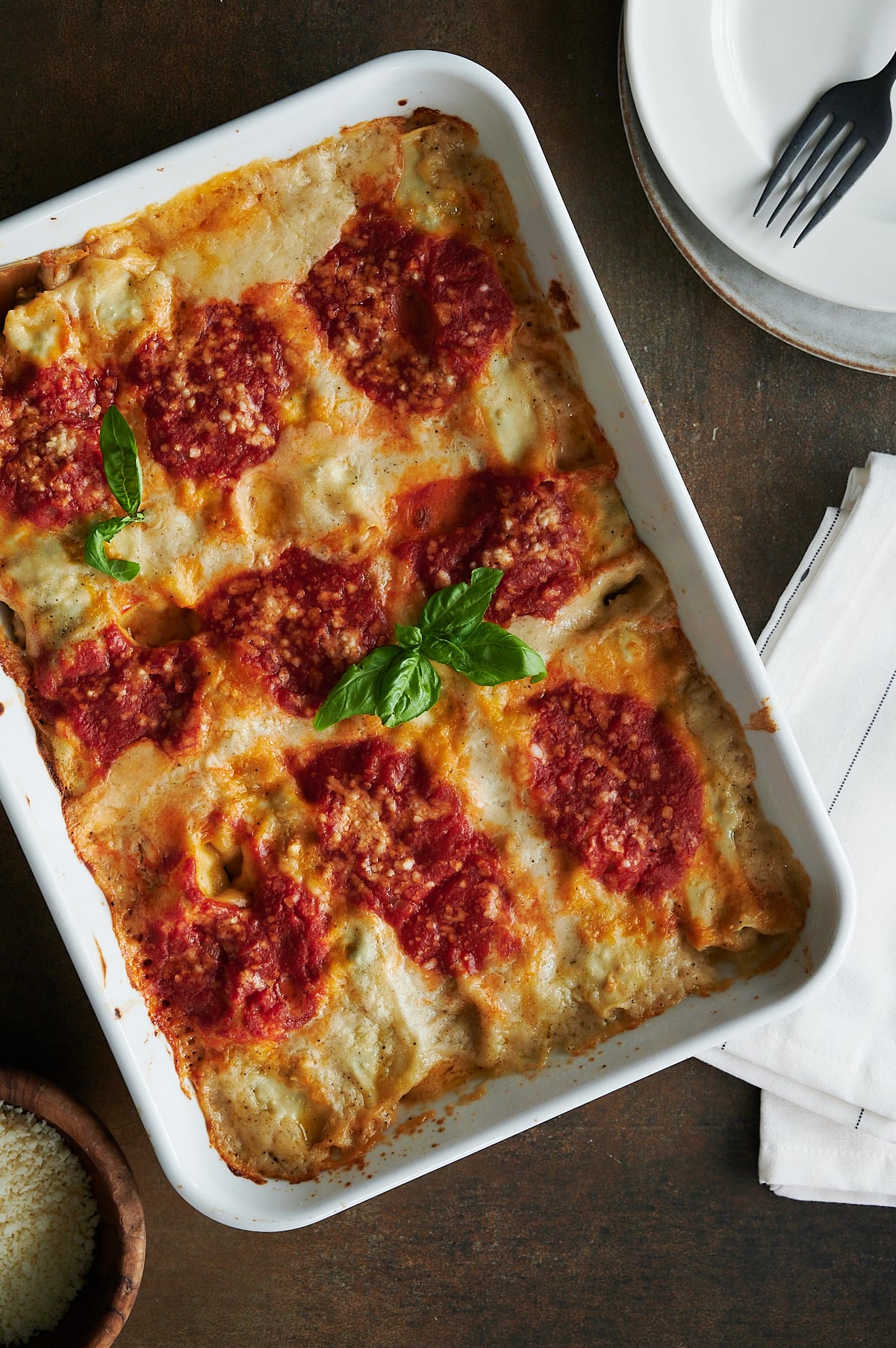 A baked lasagna in a white dish topped with tomato sauce and fresh basil, placed on a wooden table next to a stack of plates, a fork, and a folded napkin.