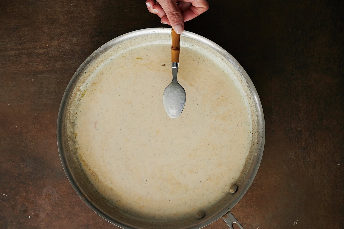 A hand holds a spoon over a large pan filled with a creamy white sauce on a brown surface.
