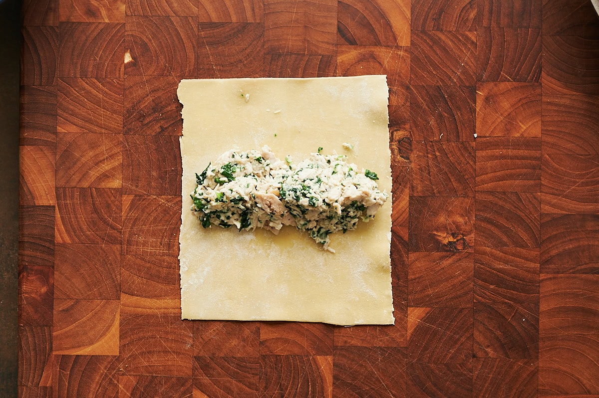 A sheet of pasta dough with a filling of chicken and spinach sits on a wooden cutting board.