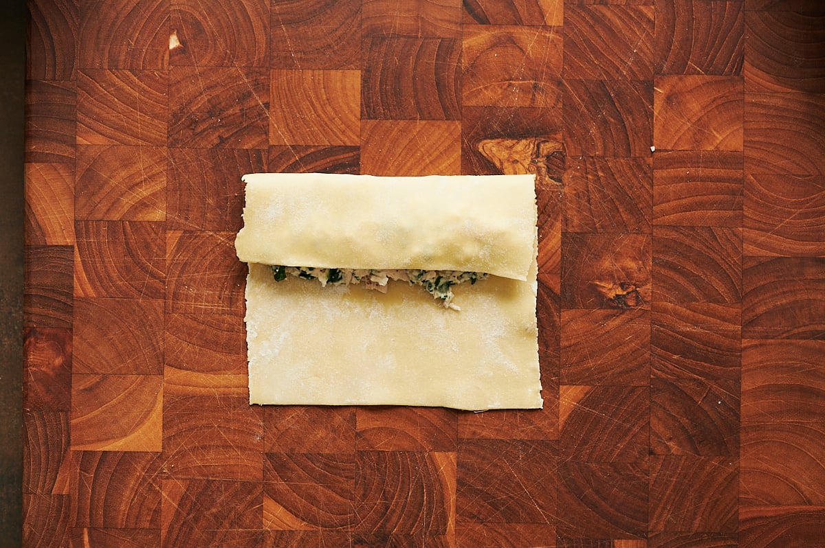 A sheet of pasta dough partially folded over a chicken and spinach filling on a wooden cutting board.