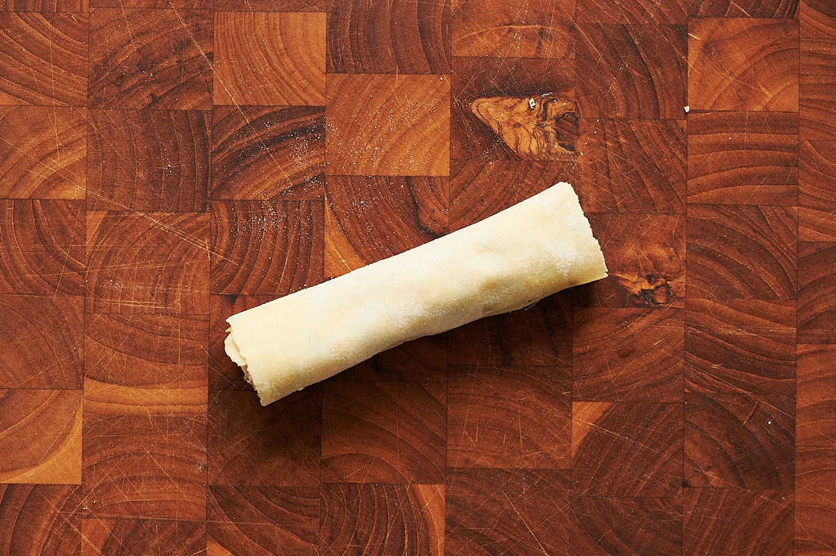 A filled, rolled and unbaked cannelloni placed on a wooden cutting board with a checkered pattern.