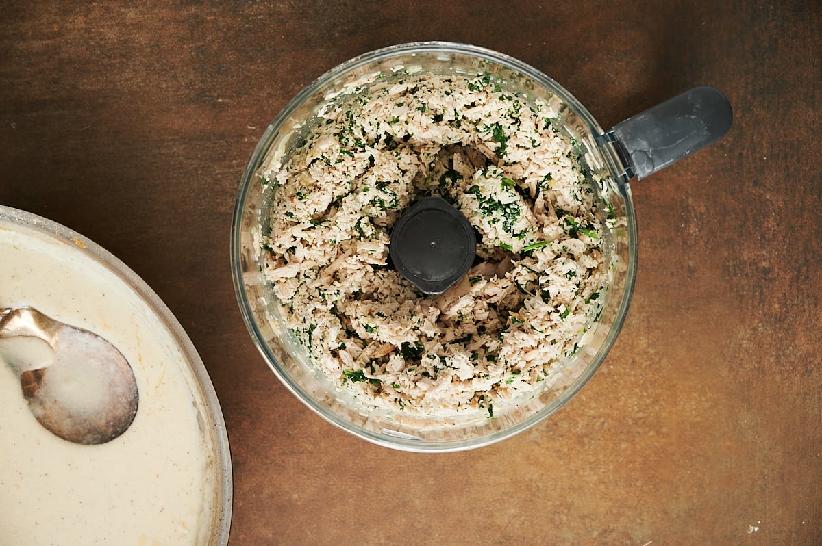 A food processor with a mixture of chopped spinach and chicken sits on a brown surface next to a bowl of white sauce with a spoon.
