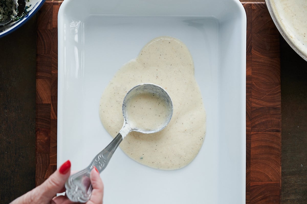 A hand pours a ladle of white sauce into a rectangular white baking dish on a wooden surface.