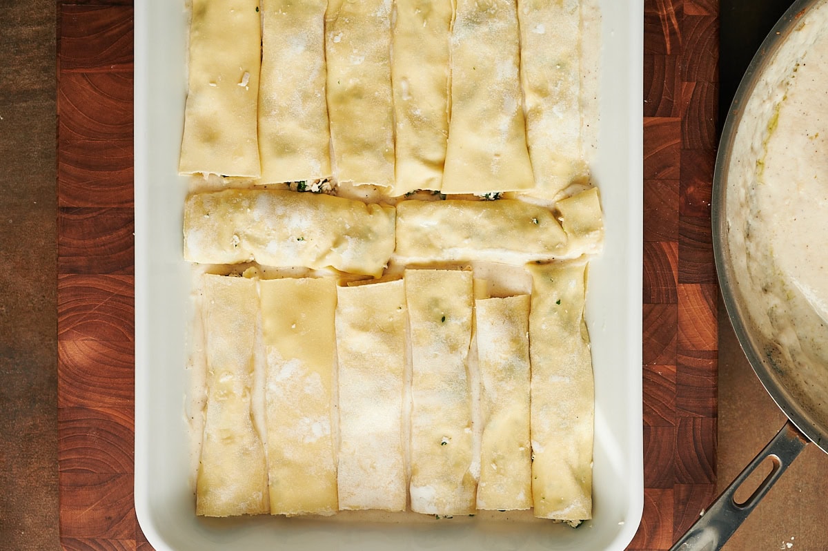 Unbaked cannelloni rolls filled with chicken and spinach are arranged in a white baking dish, ready for the oven. A pot with white sauce is partially visible on the right.