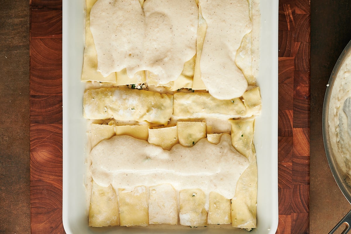 A baking dish filled with uncooked chicken cannelloni, some rolled, topped with a layer of white sauce, on a wooden surface.