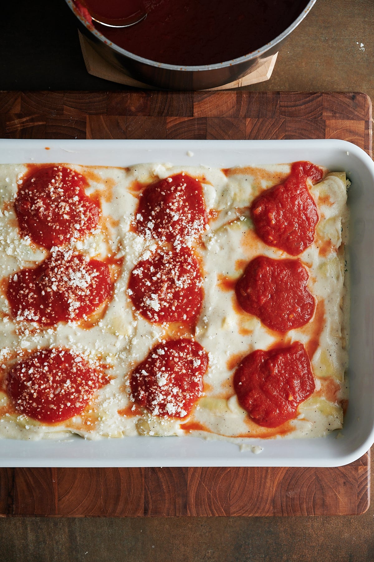 A white baking dish with partially assembled chicken cannelloni, featuring béchamel sauce, tomato sauce, and grated cheese sprinkled on top.